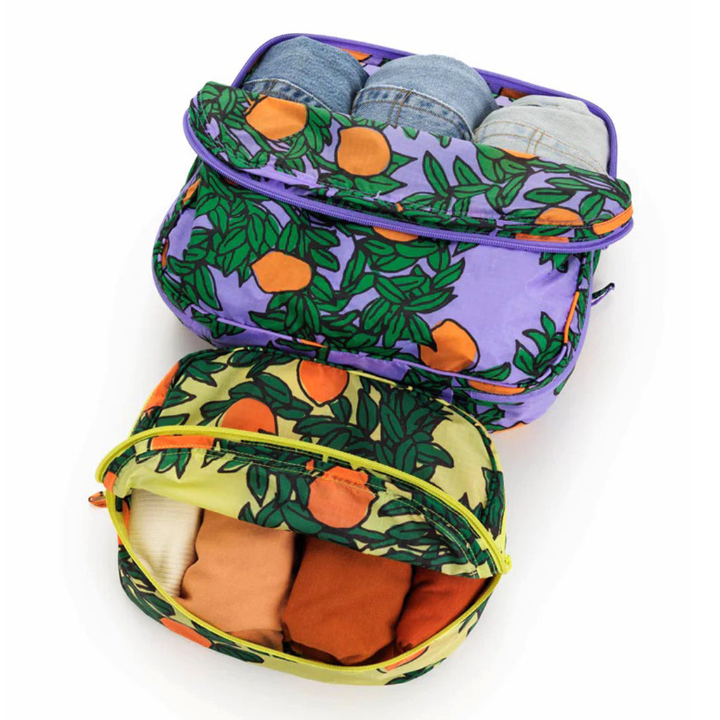 Baggu reusable recycled ripstop nylon packing cube set of 2 with orange trees print, large has a periwinkle backdrop and the small cube has a yellow backdrop, partially unzipped and filled with clothing.