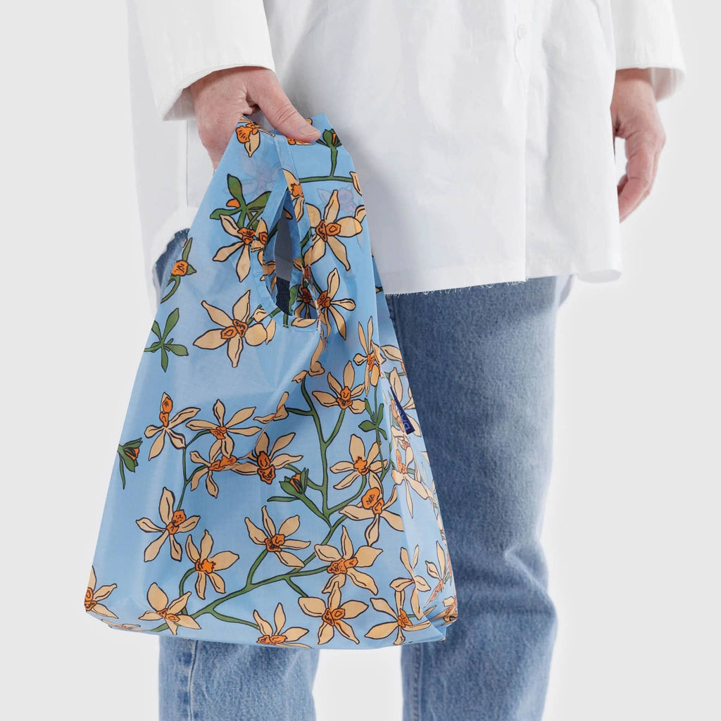 Baggu baby size eco-friendly recycled ripstop nylon reusable tote bag with the orange Orchid print on a medium blue background, in model's hand.