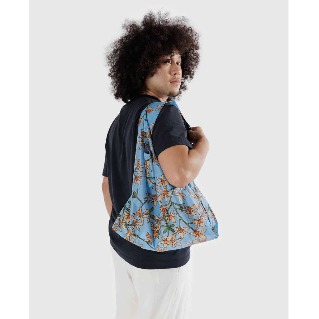 Baggu standard size eco-friendly recycled ripstop nylon reusable tote bag with an orange orchid print on a blue backdrop, on model's shoulder.