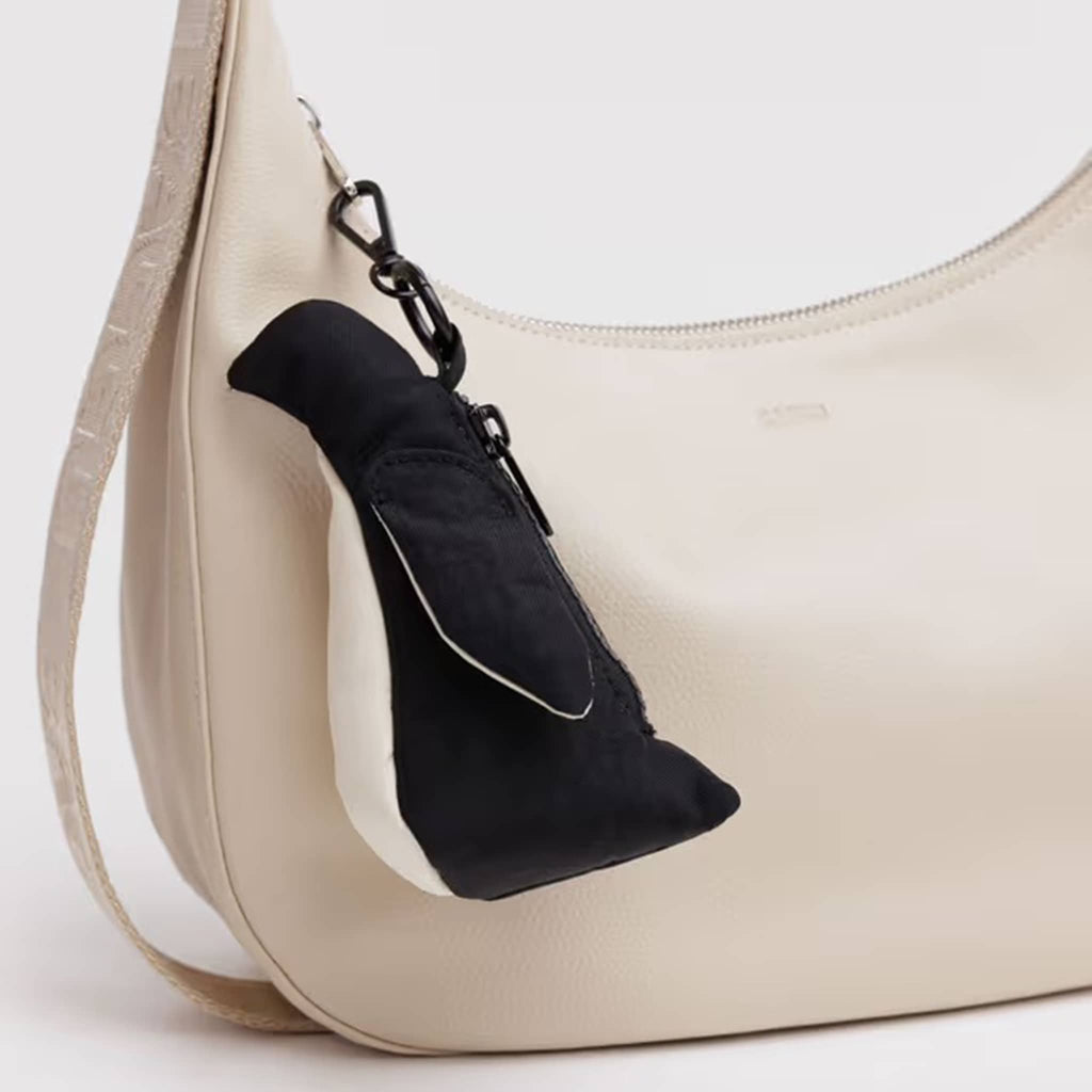 Baggu Penguin bag charm in black and white recycled nylon, clipped on a cream leather crescent bag.