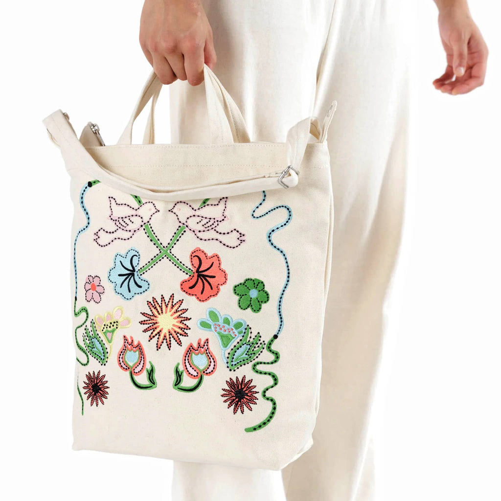 Baggu recycled cotton canvas duck bag tote with zip top closure in a cream color with colorful embroidered birds and flowers, in models hand.
