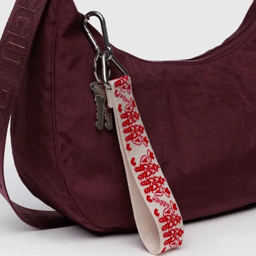 A white webbed nylon keychain with the Cowgirl print in red with Baggu logo, featuring a metal clip and a triangle ring, clipped to a burgundy Baggu Crescent Bag.