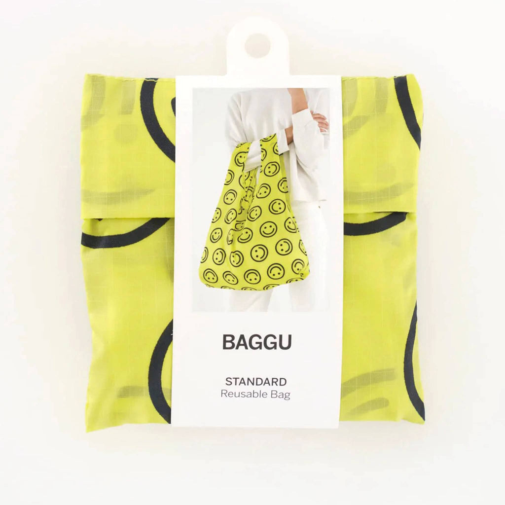 Bright yellow Baggu standard bag with repeating black smiley faces, folded into carry pouch with belly band packaging.