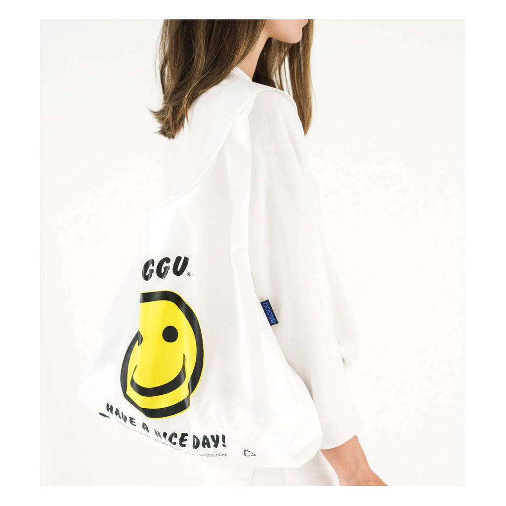 Baggu reusable standard white ripstop nylon bag in thank you happy with yellow smiley face and have a nice day in black text, on model's shoulder.