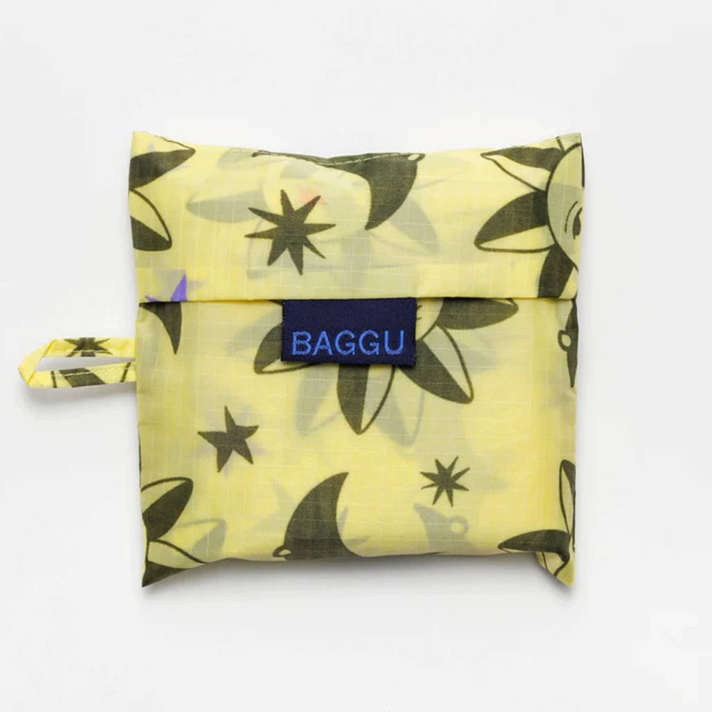Baggu standard size eco-friendly recycled ripstop nylon reusable tote bag with a sun and moon charms print on a yellow backdrop, in matching pouch.