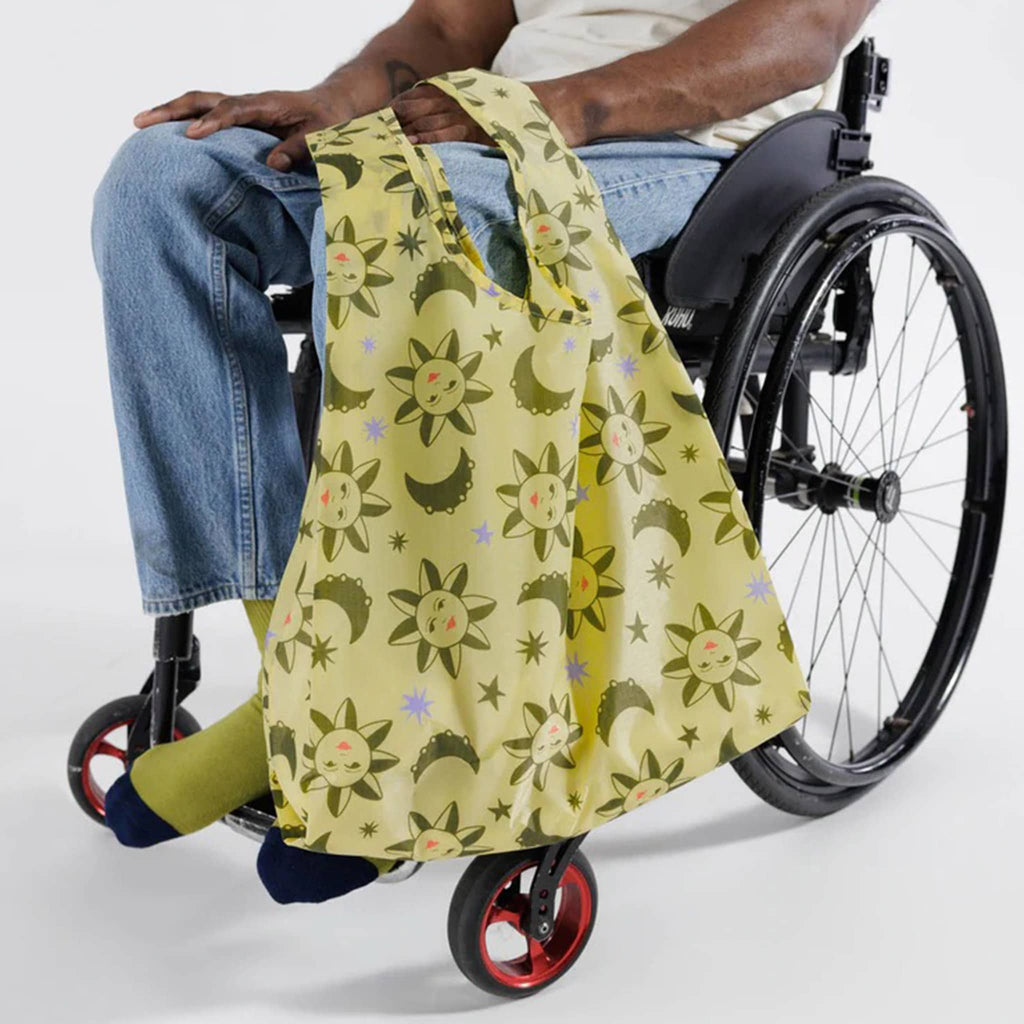 Baggu standard size eco-friendly recycled ripstop nylon reusable tote bag with a sun and moon charms print on a yellow backdrop, being held by a person in a wheelchair.