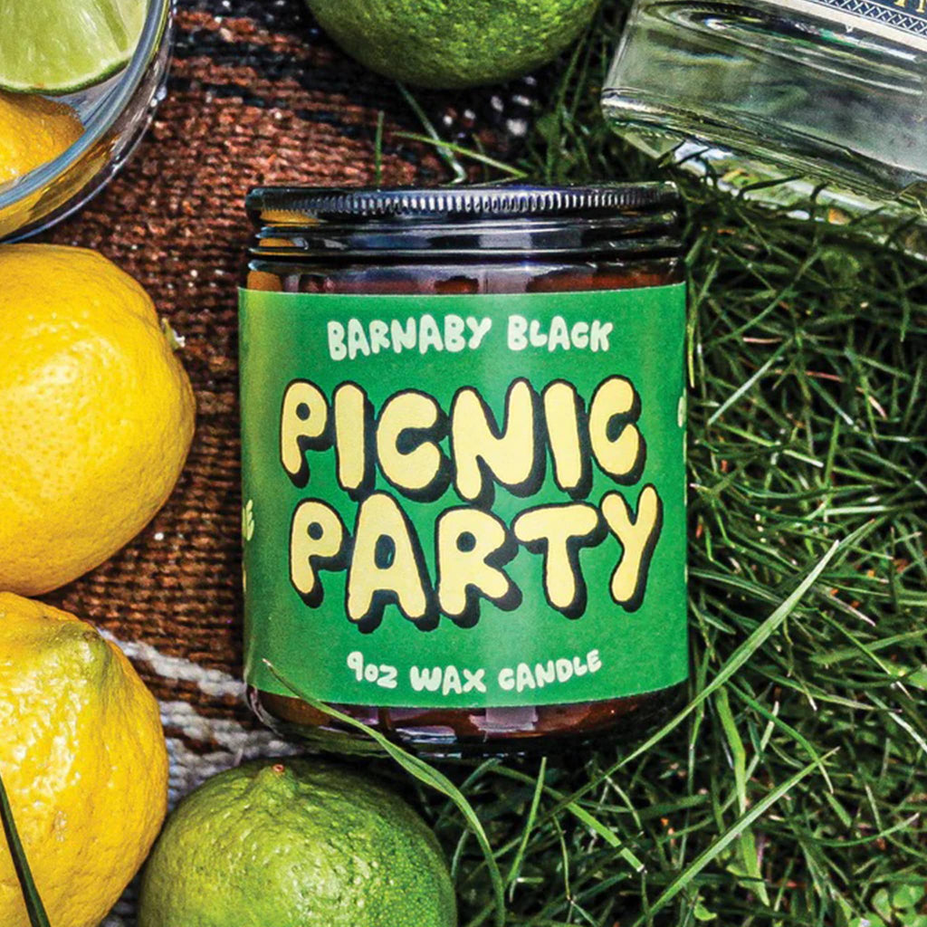 Jar of 'Picnic Party' candle by Barnaby Black surrounded by lemons and limes on a grassy background.