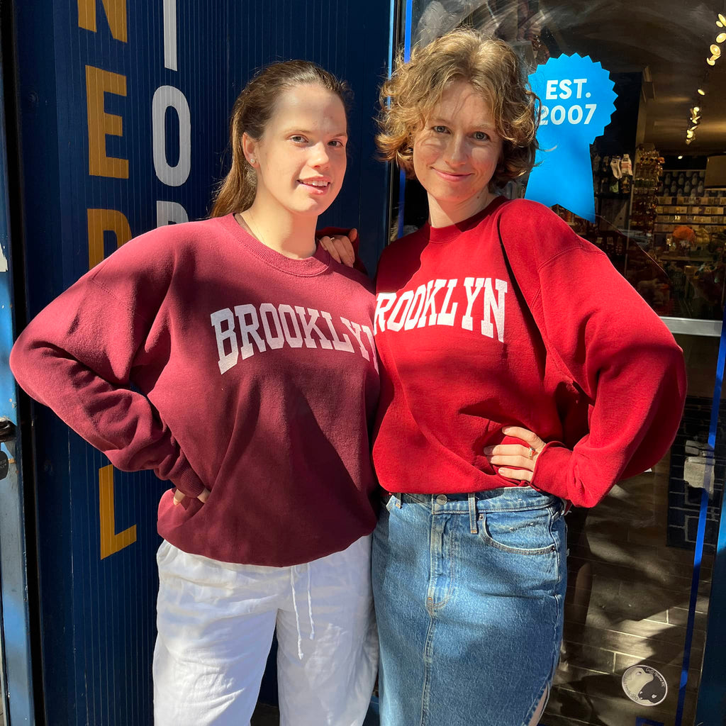 Two people wearing 'Brooklyn' sweatshirts standing in front of a blue wall with text.