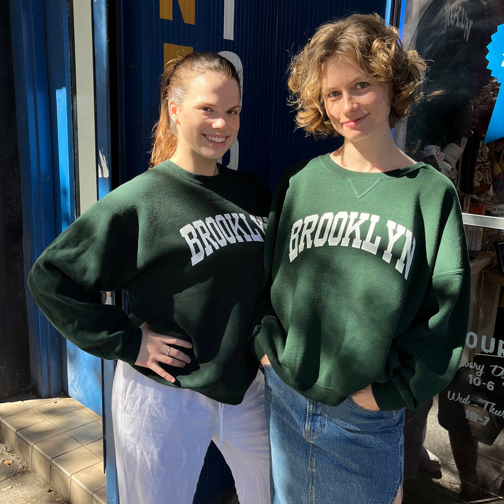 Two people wearing green 'Brooklyn' sweatshirts standing in front of a store.