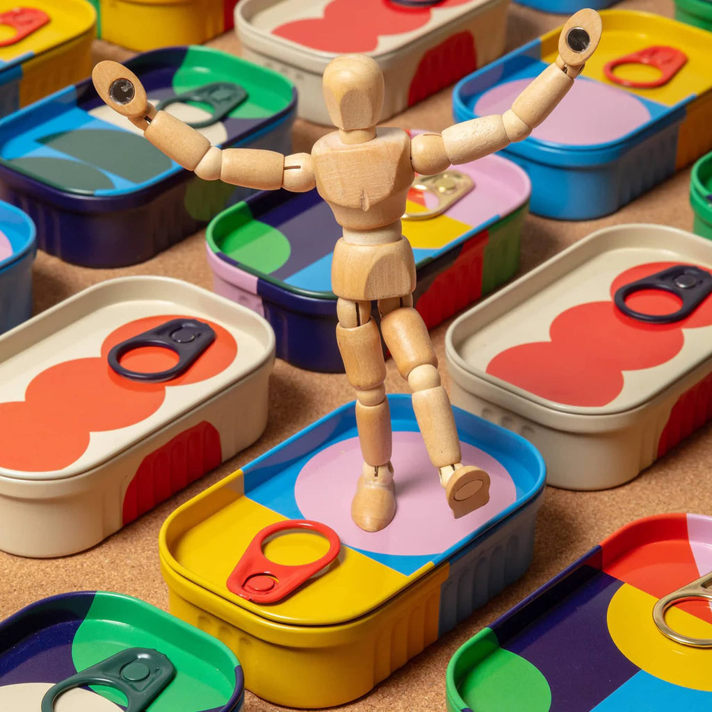 Mini wooden mannequin among colorful tins with various shapes and colors on a cork surface.