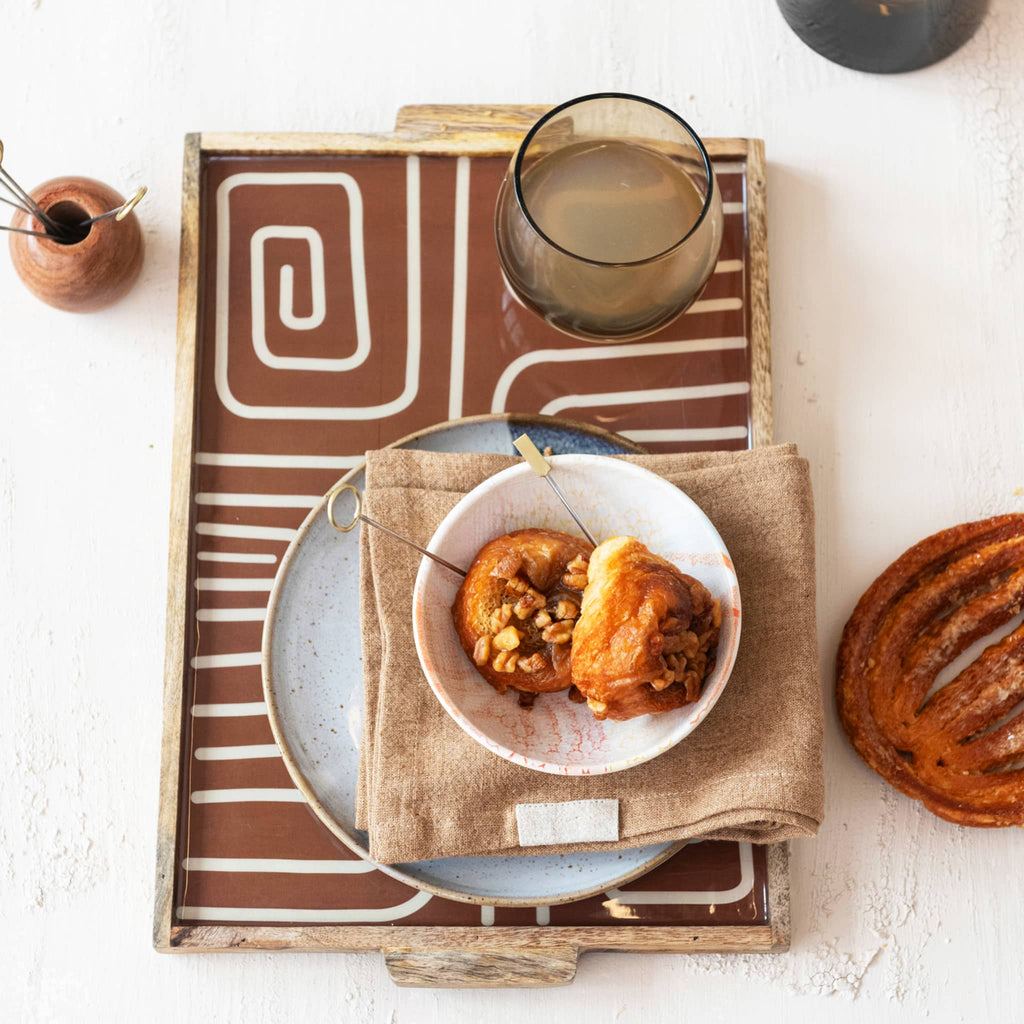 Decorative enameled wood serving tray with brown and beige abstract pattern with pecan rolls and a glass of water.