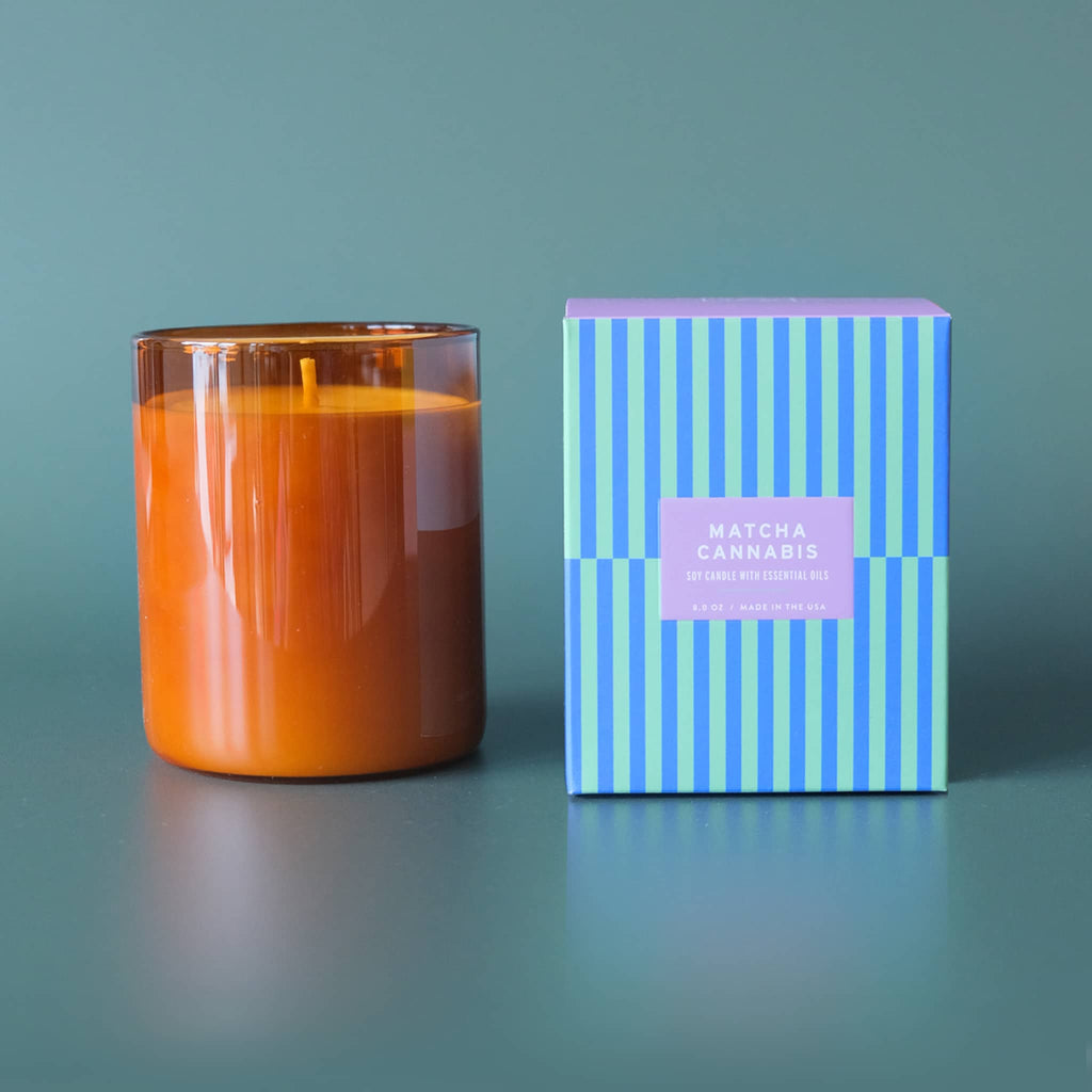 Botanica Essentials 8 ounce Matcha and Cannabyss scented soy candle with essential oils in amber glass vessel with striped box packaging.