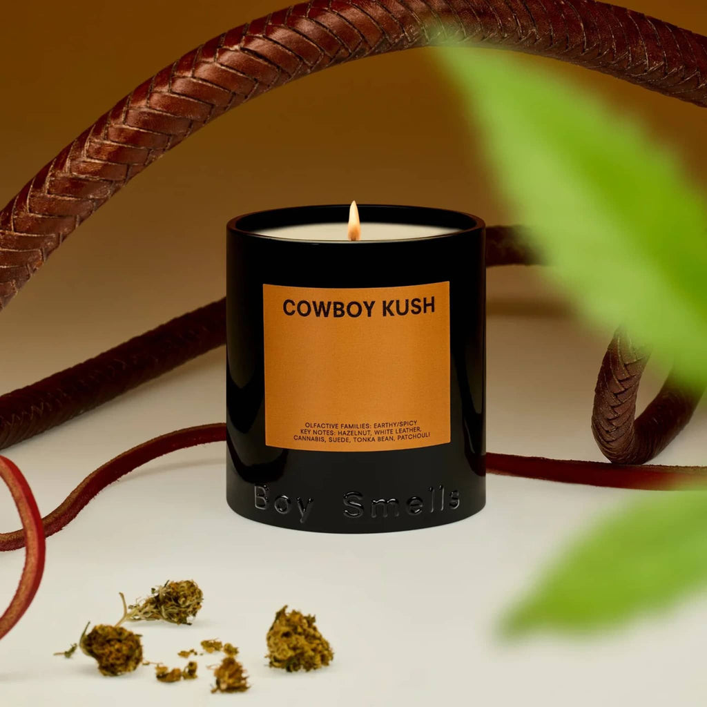 Boy Smells Cowboy Kush scented 9 ounce Classics candle in black glass vessel with dark orange label, lit with leather strips in the background.