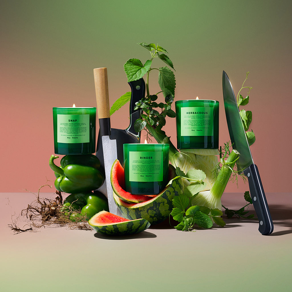 Group shot of the Boy Smells Farm to Candle Collection, all are in painted green glass tumblers and lit. Surrounded by fruits, vegetables and herbs that represent their scent, along with assorted knives.