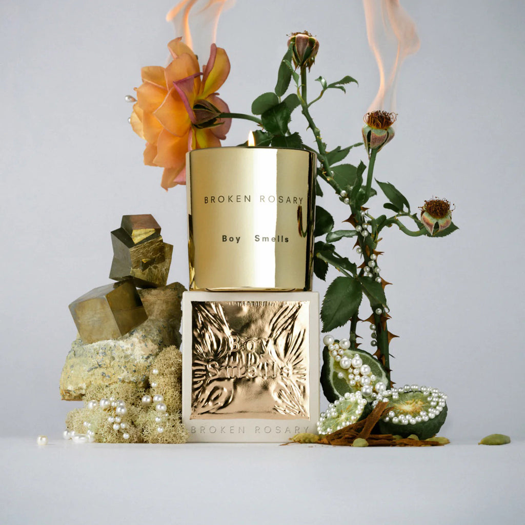 Boy Smells 2023 Limited Edition Holiday Broken Rosary scented coconut beeswax blend candle in metallic gold glass tumbler with box packaging. The candle is lit and it's on a gray background with smoking rose buds, lime, fools gold pieces and pearl beads.