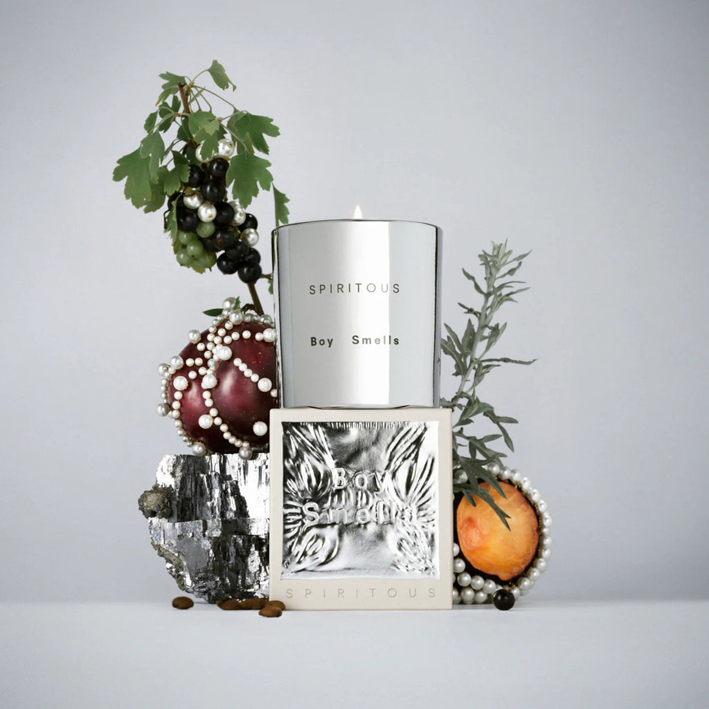 Boy Smells 2023 Limited Edition Holiday Spiritous scented coconut beeswax blend candle in metallic silver glass tumbler with box packaging. Candle is lit and it's on a gray background with fruit, leaves and berries.