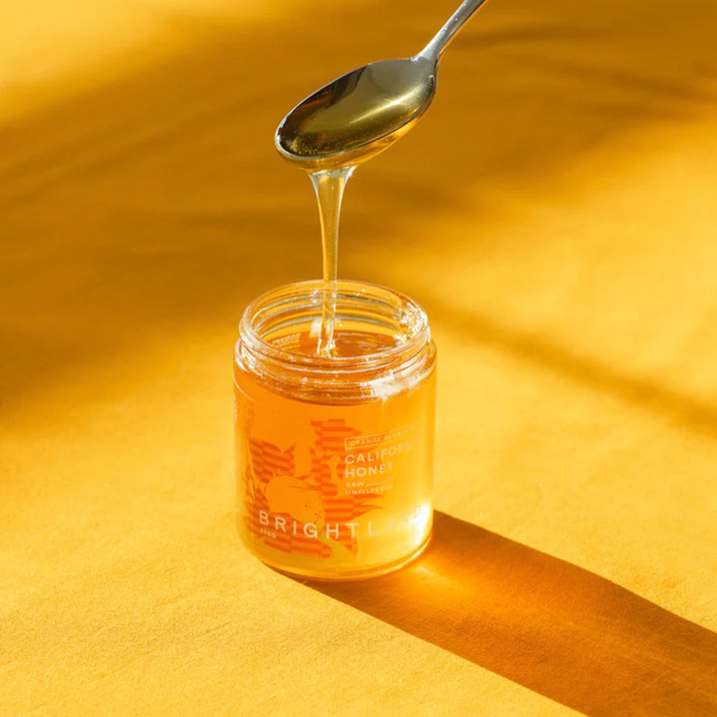 Brightland Raw & Unfiltered California Orange Blossom Honey in glass jar, a spoon is drizzling honey back into the jar.