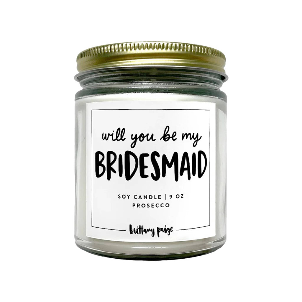 Brittany Paige prosecco scented 9 ounce soy wax candle with "will you be my bridesmaid" on the front of the clear glass jar with gold tone metal screw-on lid.