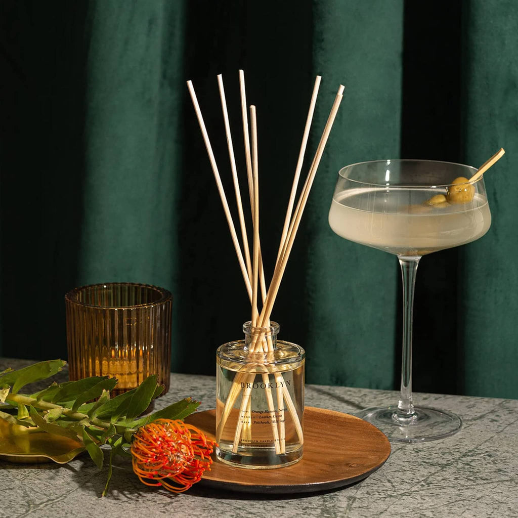 Brooklyn Candle Studio Brooklyn scented reed diffuser room perfume, clear glass bottle with reeds with martini, candle and flower with green curtain background.