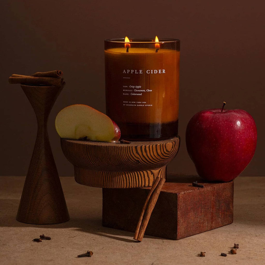 Candle labeled 'Apple Cider' with apples and cinnamon sticks on a wooden stand against a brown background.