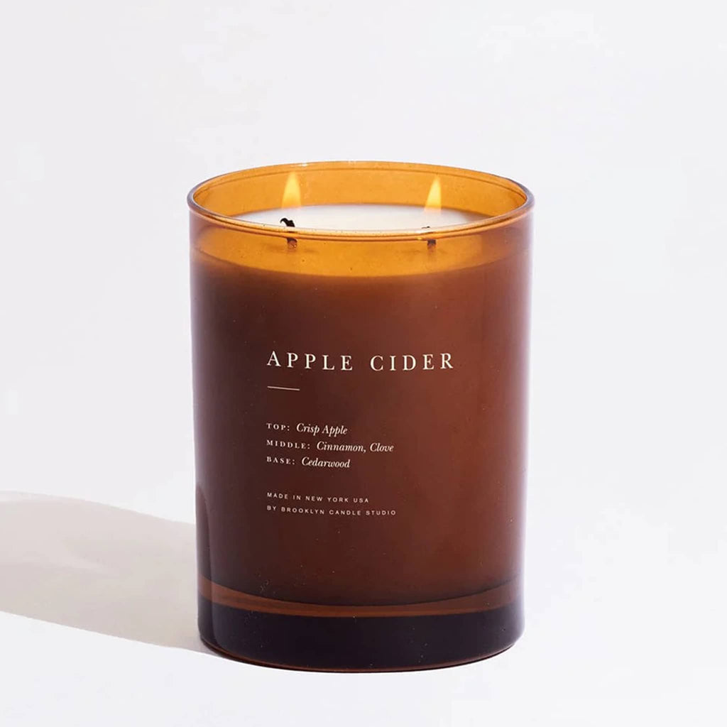 Apple cider candle with in an amber glass container, two wicks lit, on a white background.