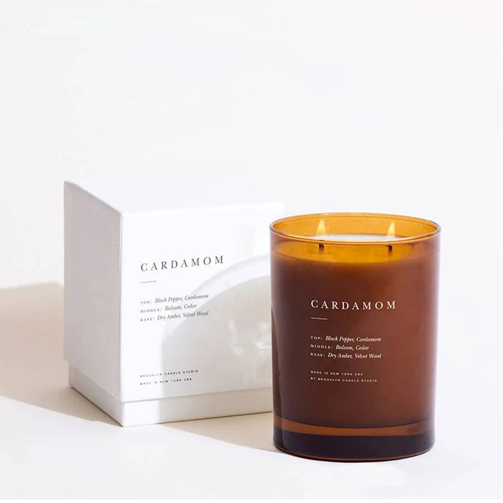 Amber glass candle vessel with 'Cardamom' label next to a white box on a light background.