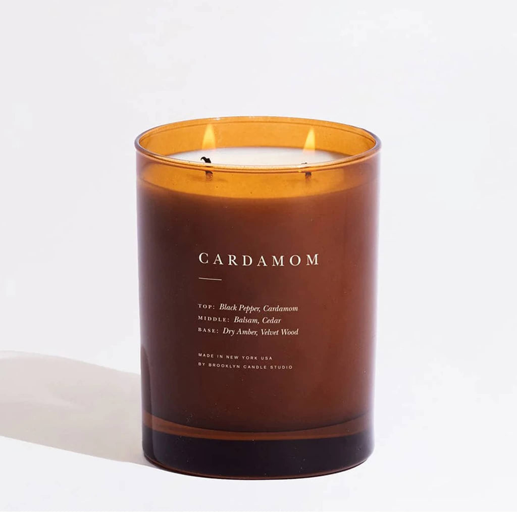 Amber glass candle vessel with 'Cardamom' label, two wicks lit, on a white background.