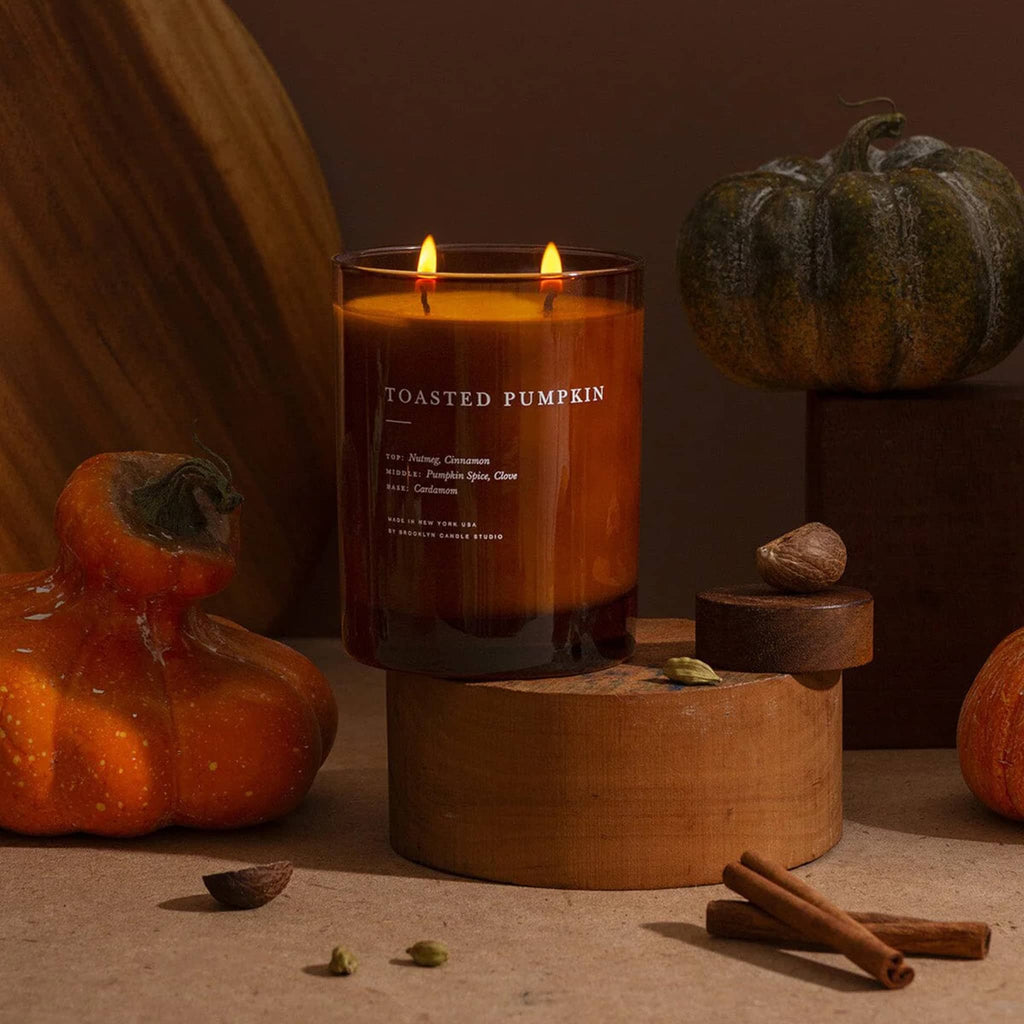 Candle labeled 'Toasted Pumpkin' surrounded by pumpkins and cinnamon sticks on a wooden surface.