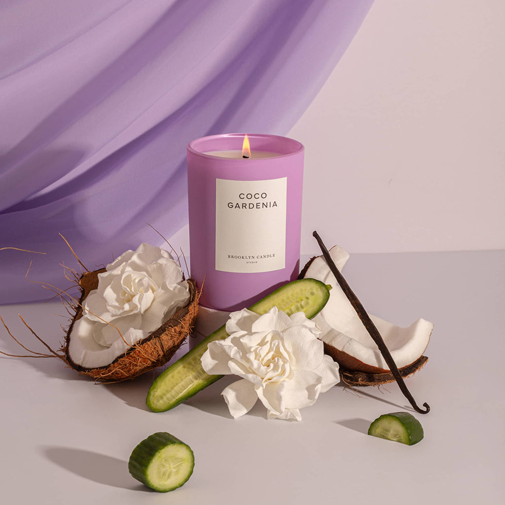 Brooklyn Candle Studio Coco Gardenia scented candle in matte lilac glass vessel with white label, lit, with coconut halves, cucumber and white gardenia blooms with purple draping in background.