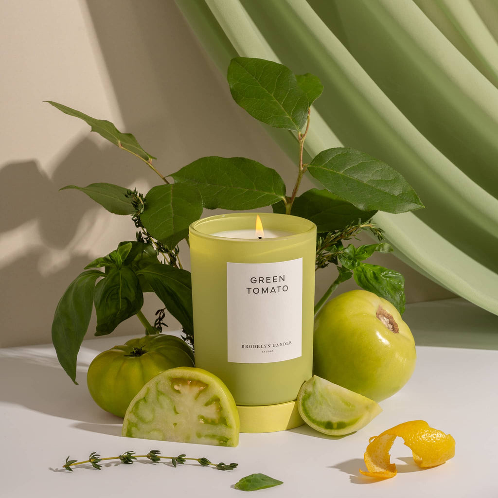 Brooklyn Candle Studio Limited Edition Herbarium Summer Collection, Green Tomato scented soy wax candle in chartreuse glass vessel surrounded by cut tomatoes with green drape behind.