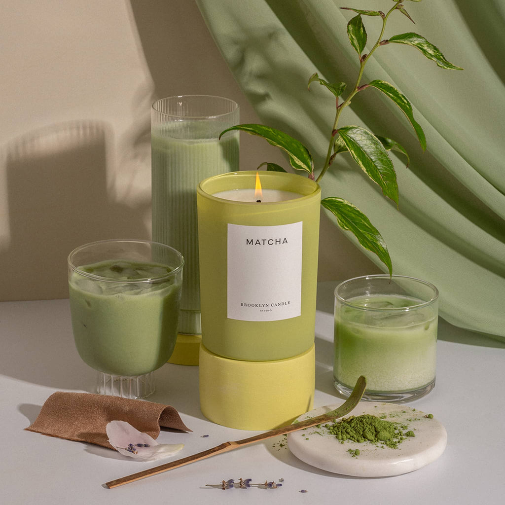 Brooklyn Candle Studio Limited Edition Herbarium Summer Collection, Matcha scented soy wax candle in chartreuse glass vessel with glasses of iced matcha and a green drape in the background.