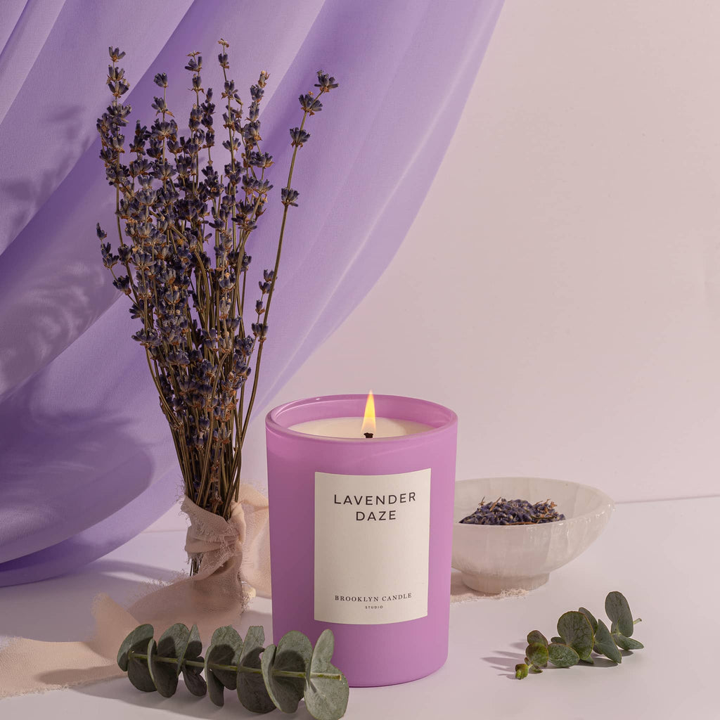 Brooklyn Candle Studio Lavender Daze scented candle in matte lilac glass vessel with white label, lit with eucalyptus leaves and lilac flower branches with purple draping in backaground.