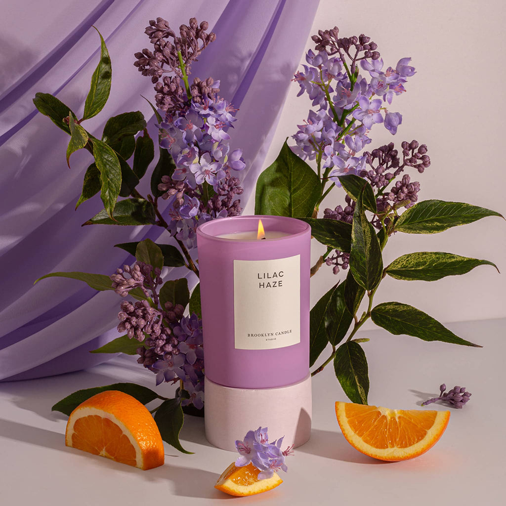 Brooklyn Candle Studio Lilac Haze scented candle in matte lilac glass vessel with white label, lit, with lilac flowers and sliced oranges with purple draping in backaground.