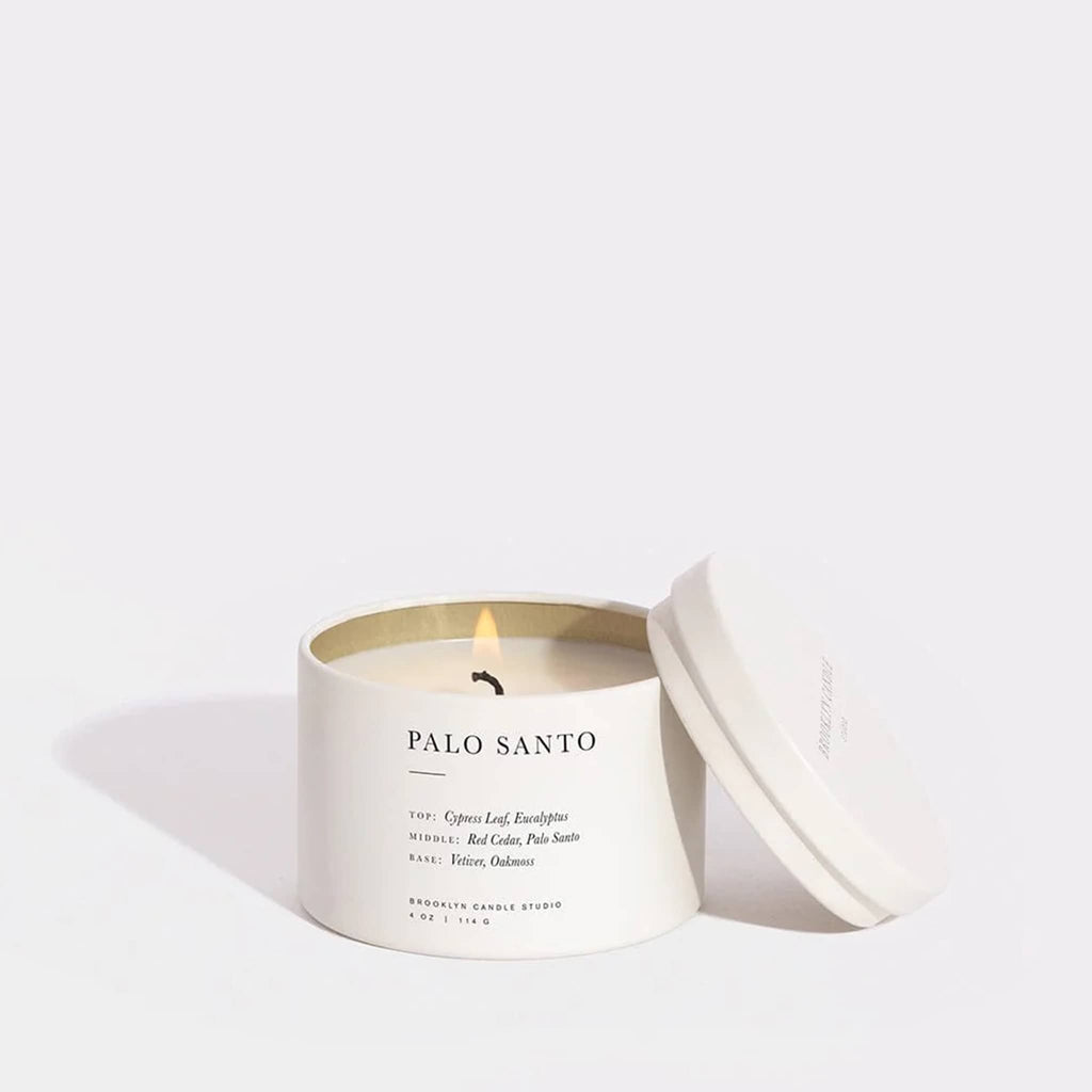 Brooklyn Candle Studio Palo Santo candle in white tin, lid off and wick lit, on a white background.