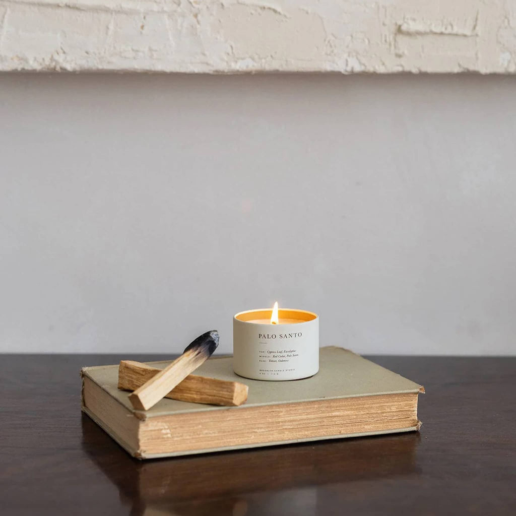 Candle labeled 'Palo Santo' on an open book with a match next to it against a neutral background.