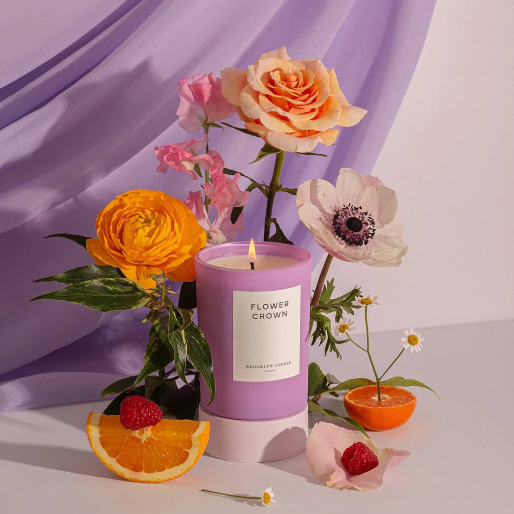 Brooklyn Candle Studio Flower Crown scented candle in matte lilac glass vessel with white label and lit wick, surrounded by flowers an orange slices with a lilac curtain sweep.