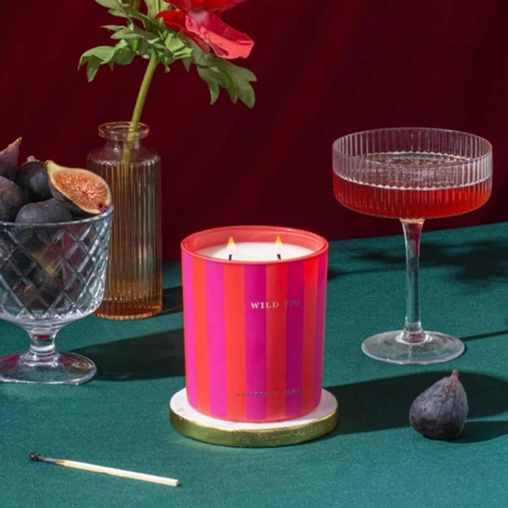 Pink and orange striped candle with 'Wild Fig' branding on a green surface with figs, a cocktail, and flowers.