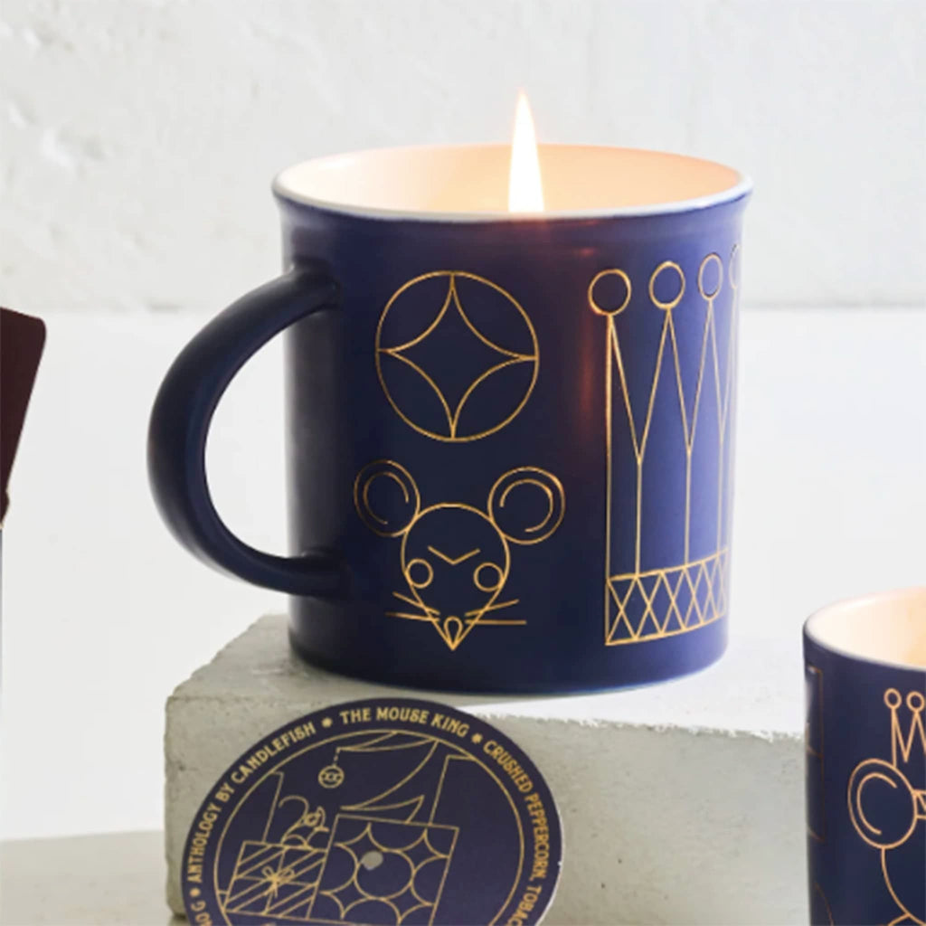 Blue mug with gold mouse king illustrations and a lit candle inside, on a light gray background.