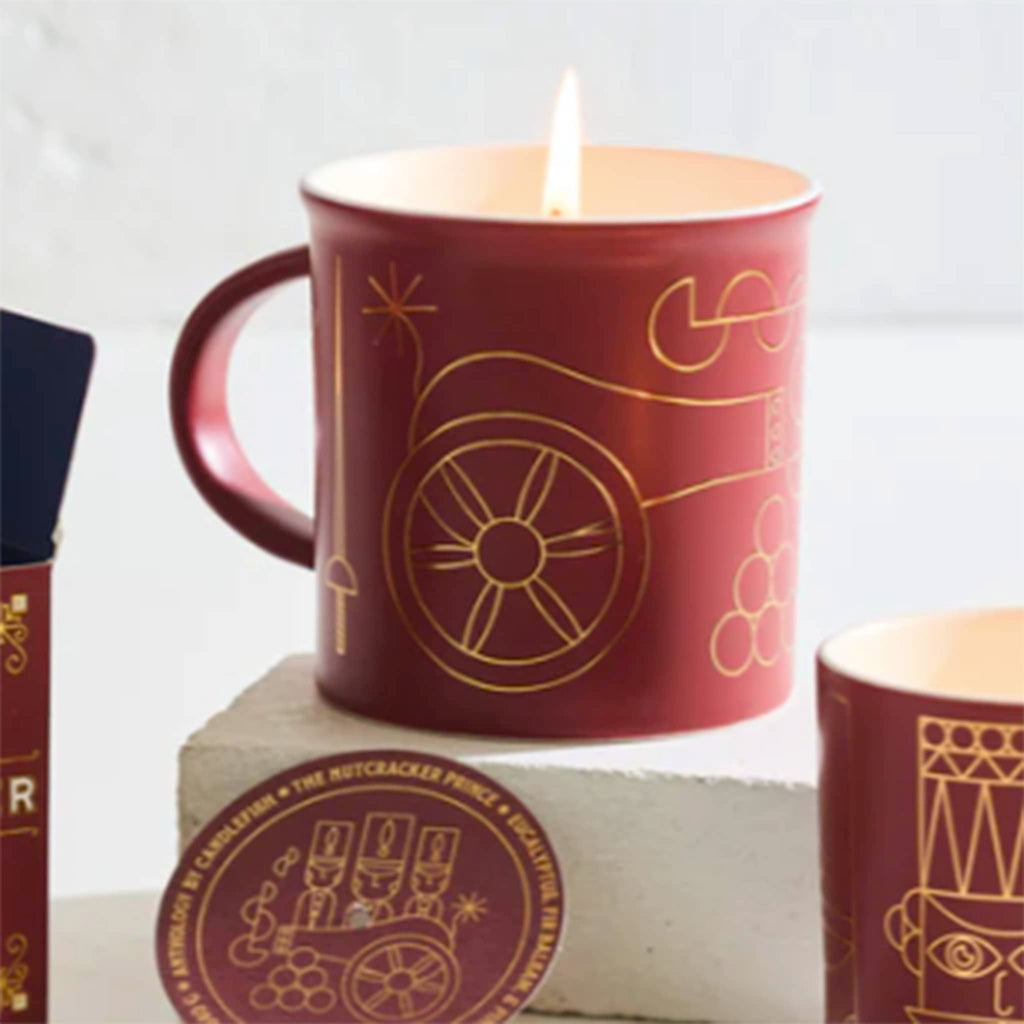 Red mug with gold nutcracker soldier illustrations and a lit candle inside, on a light gray background.