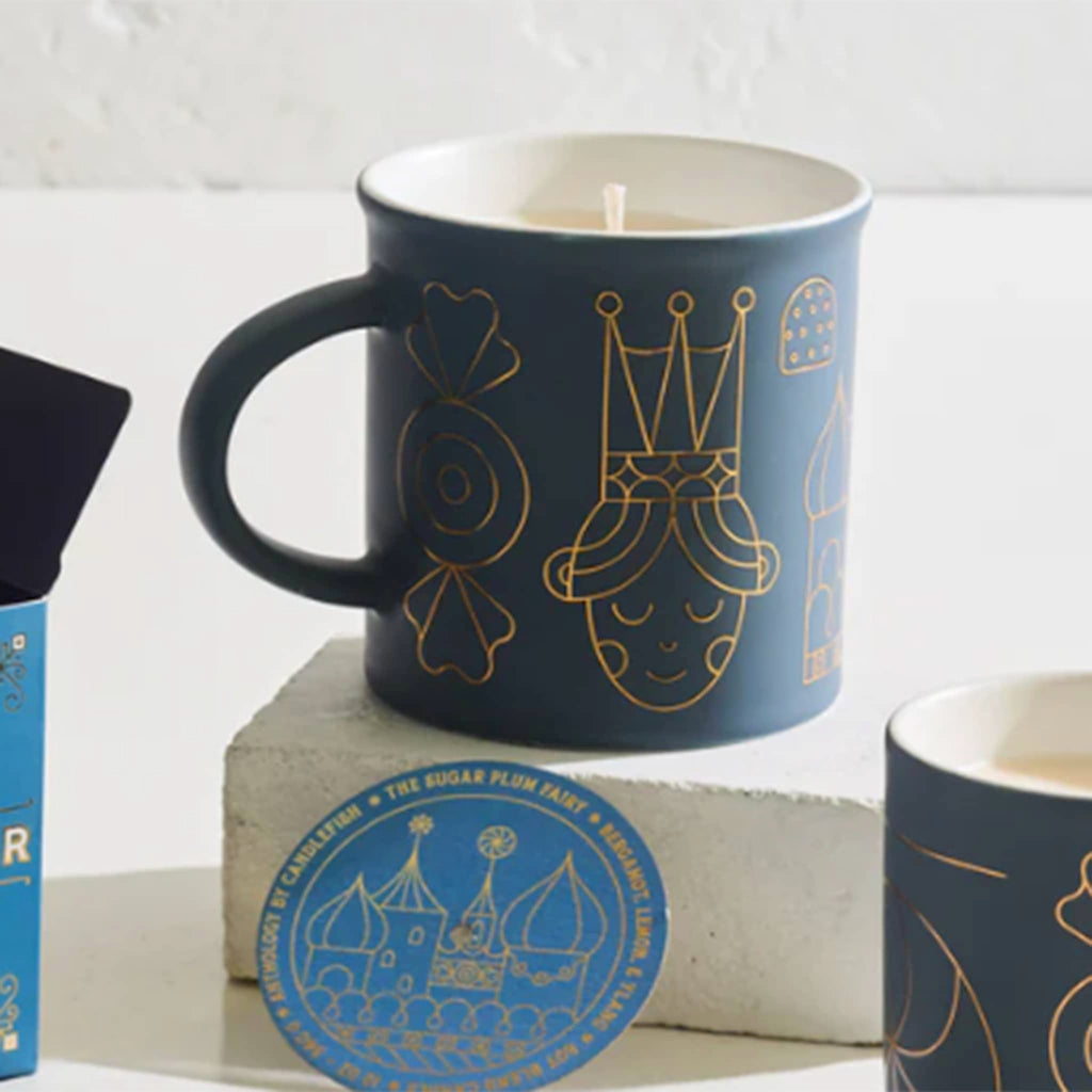 Blue mug with gold sugar plum fairy themed illustrations and a lit candle inside, on a white background.