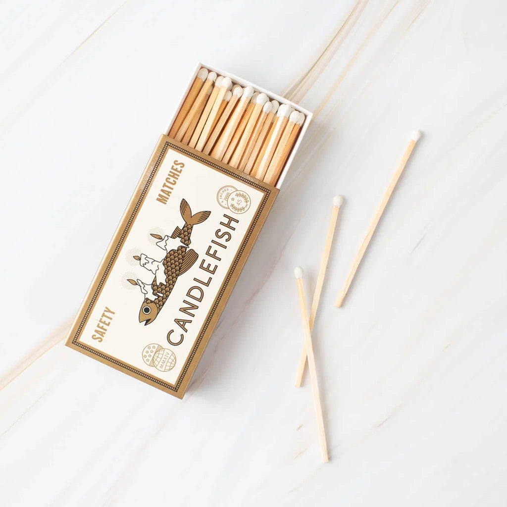 Candlefish Melting Fish matchbox with long wood safety matches with cream tips. An illustration of a fish with melting candles on its back on a cream background is shown.