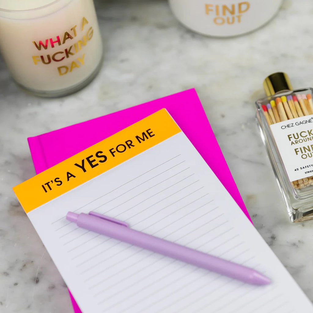 Chez Gagne ruled notepad with "it's a yes for me" in gold foil lettering on the neon orange binding at the top, sitting atop a bright pink notebook with a lilac jotter pen on it.
