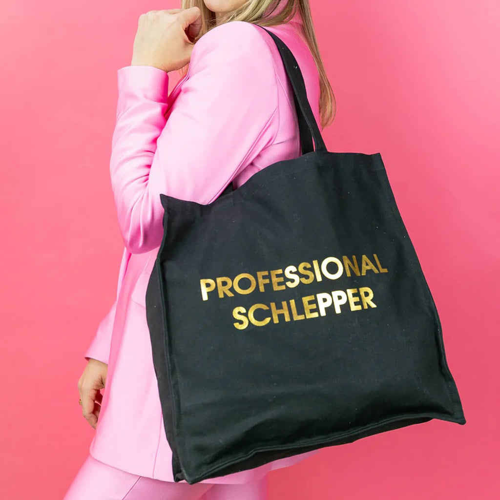 Person holding a black tote bag with 'PROFESSIONAL SCHLEPPER' in gold text against a pink background.