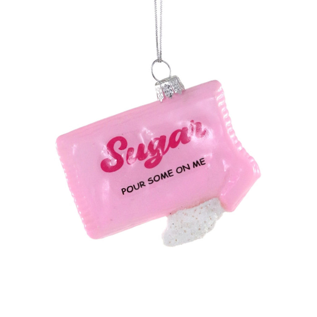 Cody Foster glass holiday tree ornament that looks like a pink sugar packet with "sugar, pour some on me" in pink and black lettering with a little "spill" of glittery sugar coming out of the bottom.