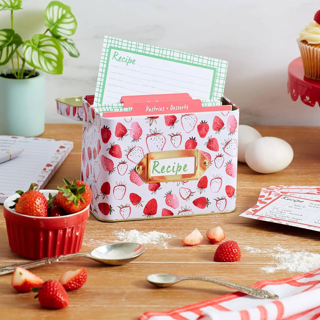 CR Gibson Strawberry Fields recipe tin box, front angle, open with recipe card coming out on a wood countertop with eggs, flour and strawberries.