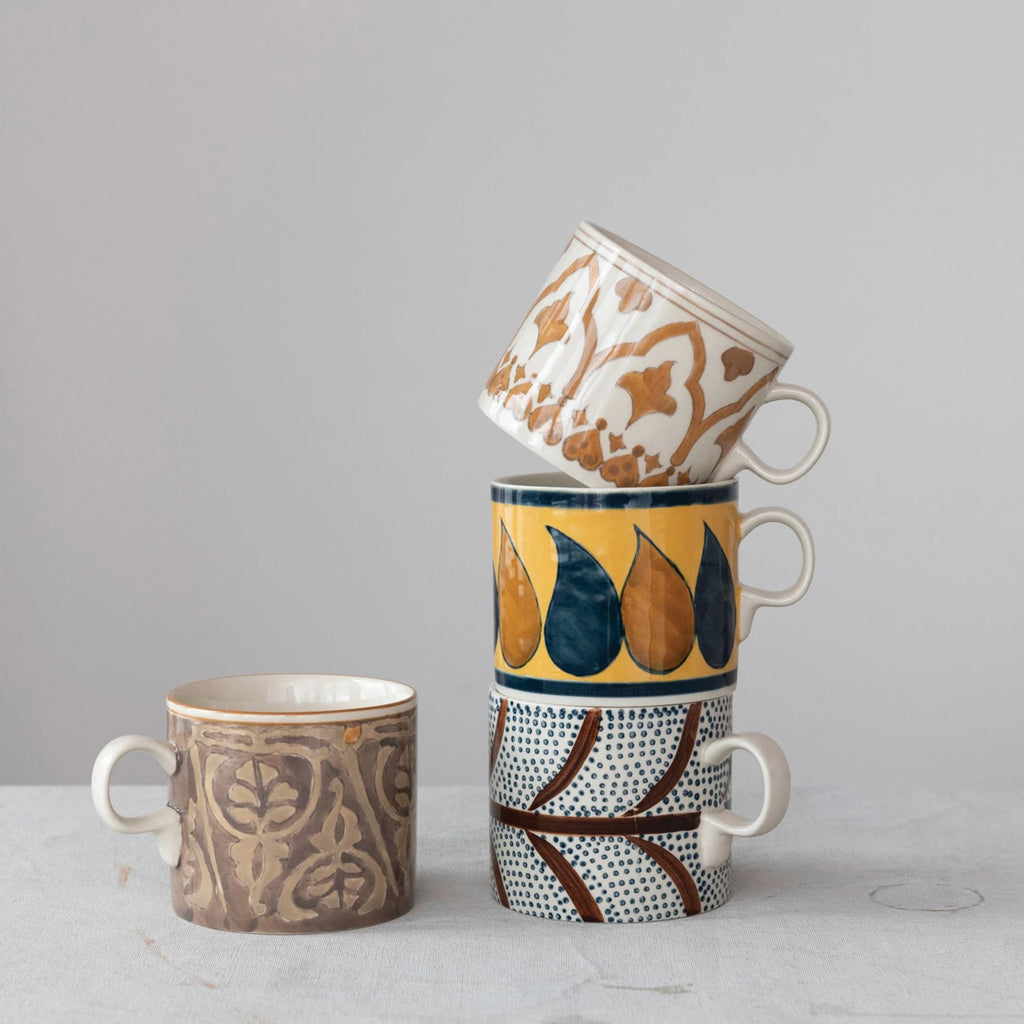 Creative Co-op 16 ounce hand-painted stoneware mugs with brown, blue, cream, tan and yellow patterns in 4 styles, 3 stacked and another beside on a gray background.