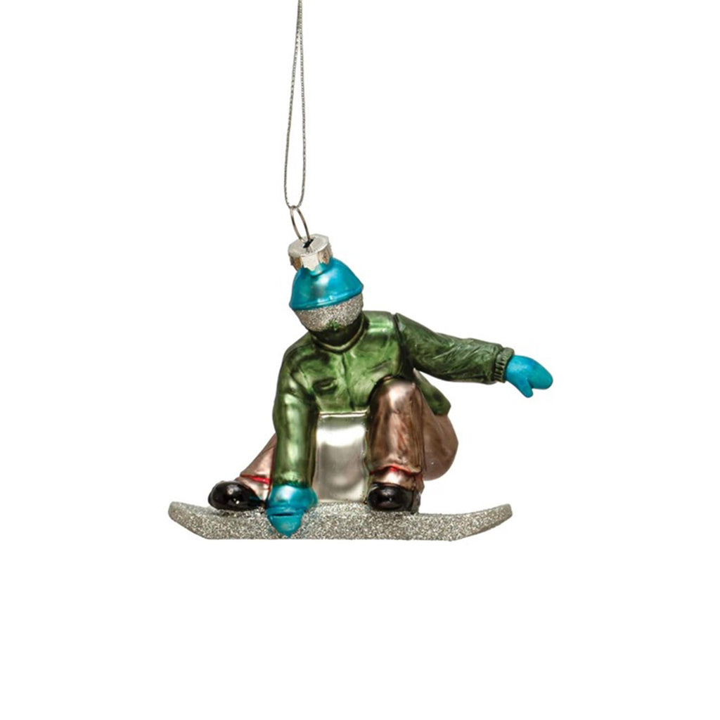 Creative Co-op snowboarder with blue hat and mittens, green jacket, tan pants and glitter silver goggles and snowboard in action hand-painted glass holiday tree ornament.
