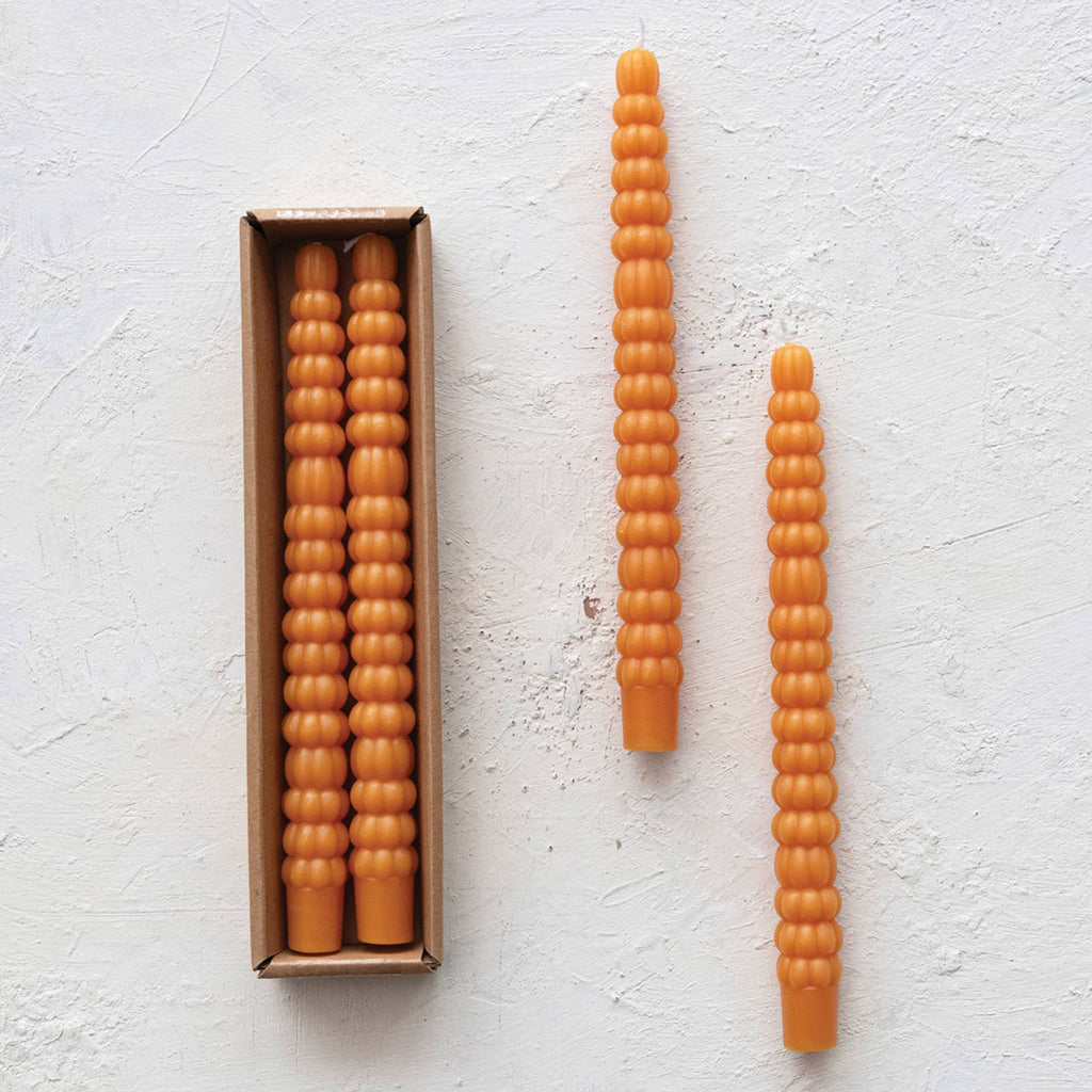 Two orange taper candles that look like stacked pumpkins in box packaging and out on a white textured surface.