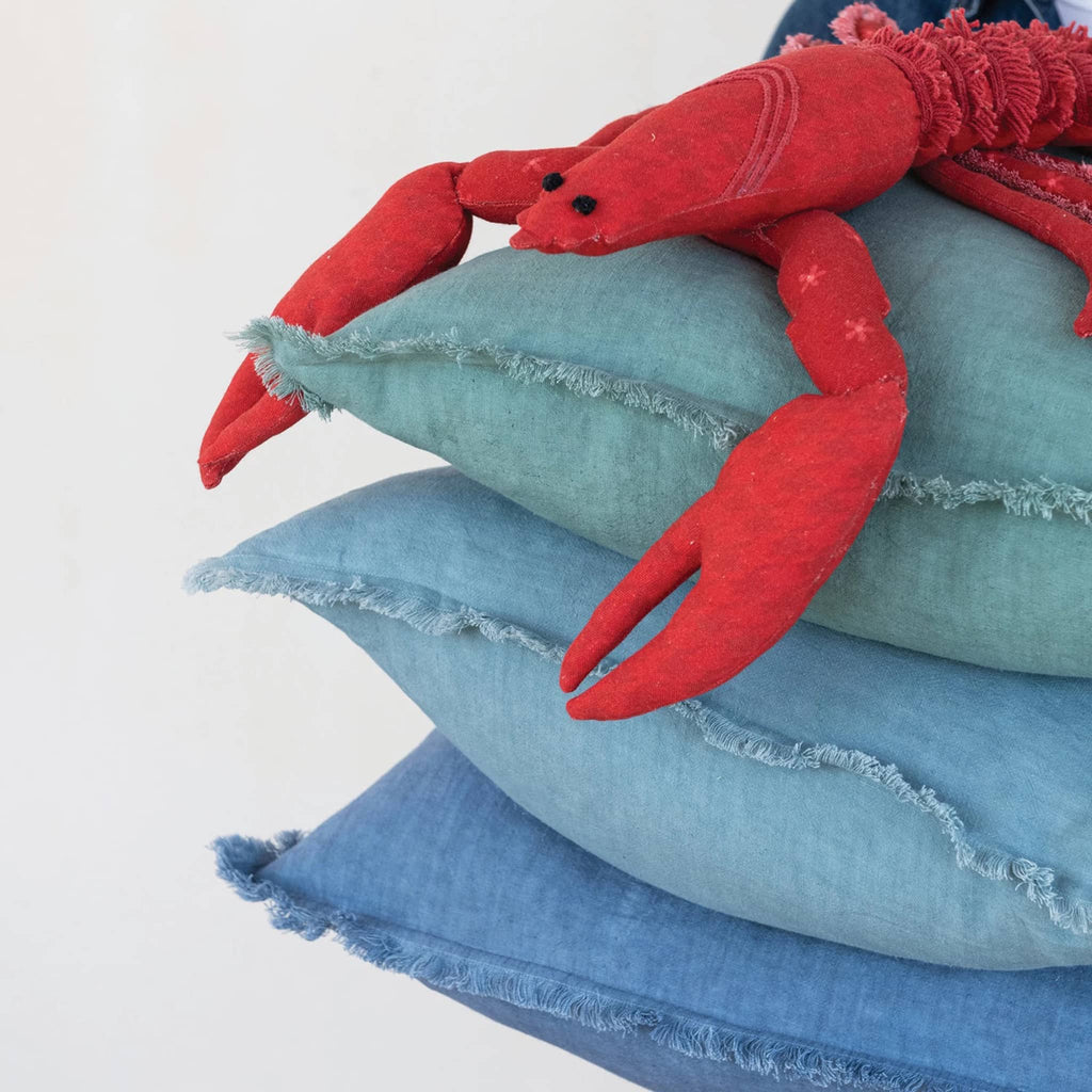 Creative Co-op cotton printed lobster-shaped pillow with embroidery and fringe, sitting on a stack of blue pillows.