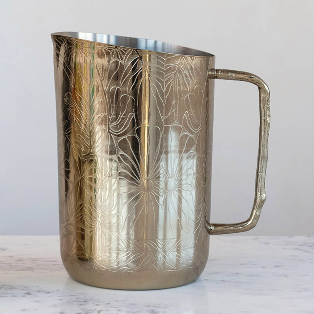 Creative Co-op Stainless Steel Pitcher with floral etched pattern and brass twig handle on the right, on a marble surface.